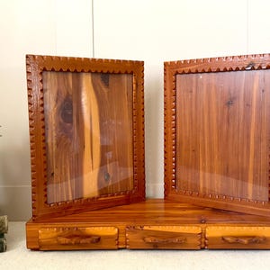 May include: Two wooden picture frames with a natural wood grain finish. The frames have a decorative, notched edge and are displayed on a matching wooden base with three small drawers. A white vase with berries is in the background.