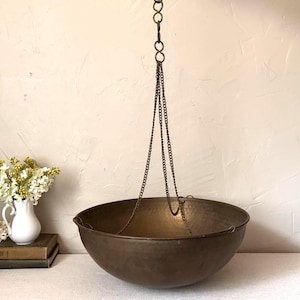 May include: A large, round, hammered bronze-colored metal bowl suspended by three thin metal chains that connect to a single vertical chain. The bowl has a wide, open top and a smooth, curved exterior surface with a dark, metallic finish.