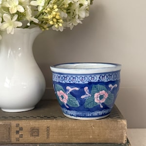 May include: A small ceramic bowl featuring a deep blue background with a repeating pattern of pink flowers and green leaves. The rim and base are decorated with a thin blue geometric border. The bowl has a smooth, glossy finish and a white interior.