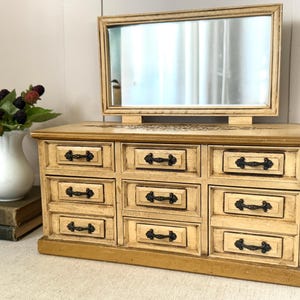 May include: A vintage-style jewelry box with a mirror, featuring a light beige finish and nine drawers with black hardware. The mirror is rectangular and framed in a matching light beige. A white vase with berries and a stack of books are in the background.