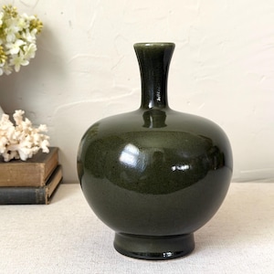 May include: A dark olive green ceramic vase with a smooth, glossy finish, featuring a rounded, bulbous body, a narrow neck, and a flared rim, standing on a circular base. The surface reflects light, highlighting the deep, uniform color.