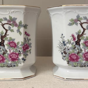 May include: A pair of white porcelain vases with an octagonal shape and gold-trimmed rims. Each vase features a colorful floral pattern depicting pink peony-like flowers, green leaves, and brown branches on a white background with embossed details.