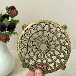 May include: A round, antique brass trivet with an openwork design. The trivet features a central hole and a repeating pattern of stylized floral shapes. The trivet is held by a hand, and a white vase with berries is in the background.