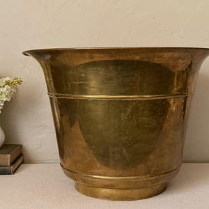 May include: A large, polished brass planter with a wide, flared rim and a tapered body. The metal surface features a warm, golden-brown patina with subtle variations in tone, a horizontal band detail, and a sturdy, circular base.