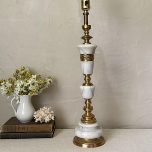 May include: A tall table lamp featuring a white marble body with polished brass accents. The lamp has a tiered circular base, a central column with alternating marble and brass sections, and a decorative brass band with an embossed floral pattern.