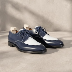 May include: A pair of two-tone derby shoes. The shoes are navy blue with white accents on the toe and a white stitch. The shoes have dark blue laces and are sitting on a light gray surface.