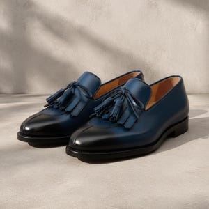 Handmade men's navy blue patina tassel loafers in genuine cowhide leather with hand-burnished indigo-to-midnight colour graduation and tonal hand-cut leather tassel, photographed at low angle on a concrete surface with cedar shoe trees — bespoke burnished slip-on formal dress loafers.