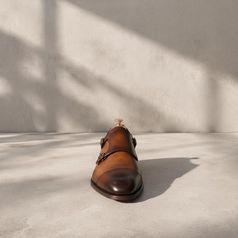Handmade men's tan brown double monk strap shoes with hand-burnished cognac-to-chocolate patina finish and gold-tone buckles, displayed on a fur surface with cedar shoe trees — bespoke cap toe formal dress shoes with hand-applied patina leather.