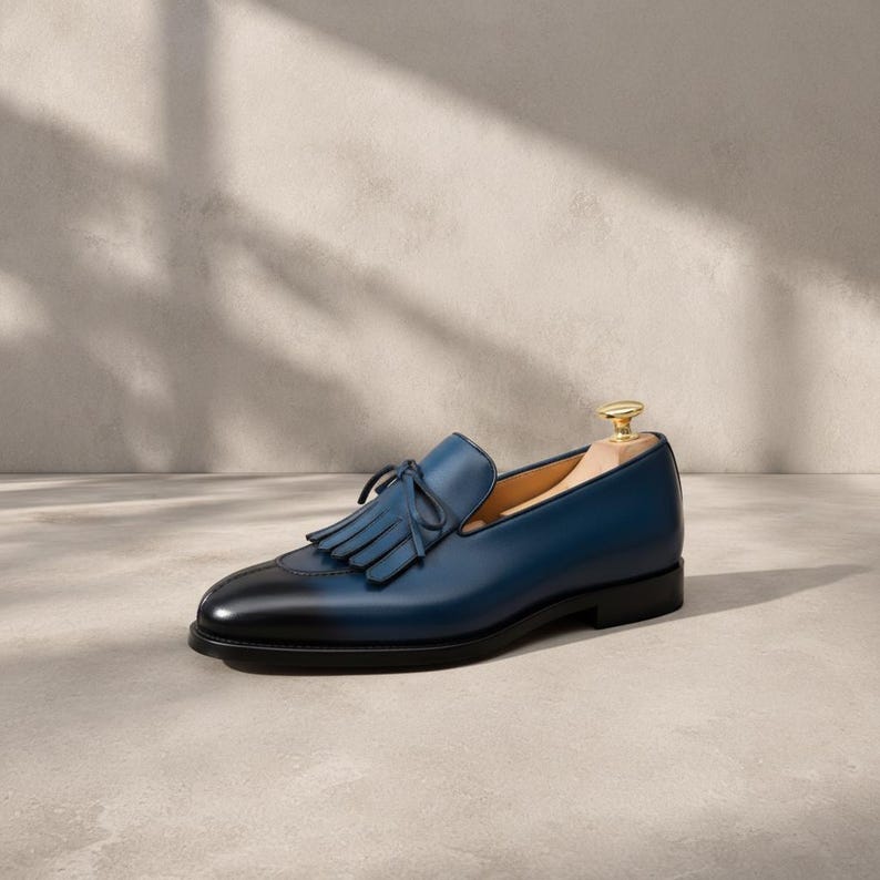 Close-up of the tonal hand-cut leather tassel with braided base and full fringe on a handmade navy patina cowhide leather loafer vamp, showing indigo-to-midnight blue burnishing graduation, smooth apron construction, and warm tan leather lining detail at the collar.