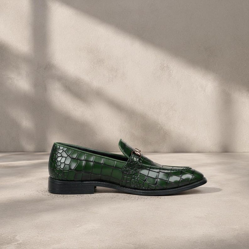 Puede incluir: Un solo mocas&iacute;n de cuero verde con textura de piel de cocodrilo. El zapato tiene un tac&oacute;n bajo y un adorno met&aacute;lico dorado en la pala. El zapato est&aacute; sobre un fondo neutro.