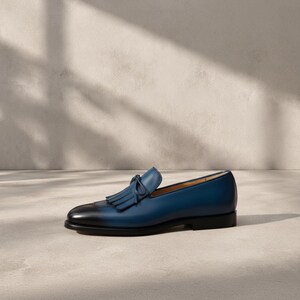 Handmade men's navy blue patina tassel loafers in genuine cowhide leather with hand-burnished indigo-to-midnight colour graduation and tonal hand-cut leather tassel, photographed at low angle on a concrete surface with cedar shoe trees — bespoke burnished slip-on formal dress loafers.