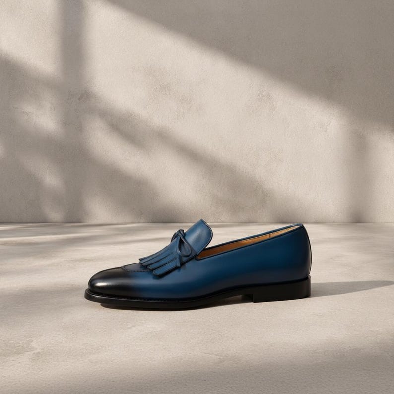 Handmade men's navy blue patina tassel loafers in genuine cowhide leather with hand-burnished indigo-to-midnight colour graduation and tonal hand-cut leather tassel, photographed at low angle on a concrete surface with cedar shoe trees — bespoke burnished slip-on formal dress loafers.