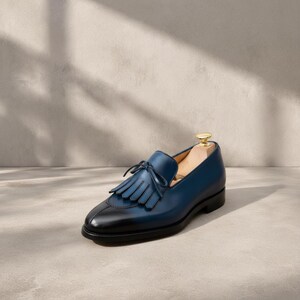 Close-up of the tonal hand-cut leather tassel with braided base and full fringe on a handmade navy patina cowhide leather loafer vamp, showing indigo-to-midnight blue burnishing graduation, smooth apron construction, and warm tan leather lining detail at the collar.