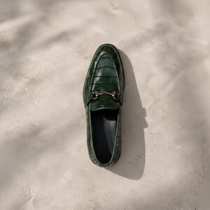 Puede incluir: Un solo mocas&iacute;n verde oscuro con textura de piel de cocodrilo. El zapato presenta una hebilla de metal dorado y una puntera redondeada. El mocas&iacute;n se encuentra sobre un fondo neutro, mostrando su dise&ntilde;o y detalles.