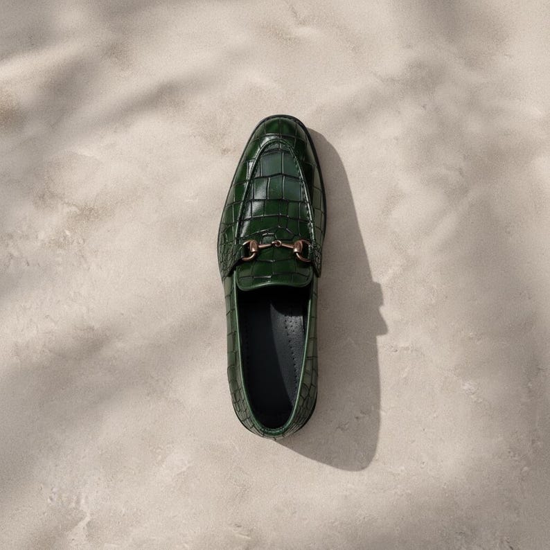 Puede incluir: Un solo mocas&iacute;n verde oscuro con textura de piel de cocodrilo. El zapato presenta una hebilla de metal dorado y una puntera redondeada. El mocas&iacute;n se encuentra sobre un fondo neutro, mostrando su dise&ntilde;o y detalles.