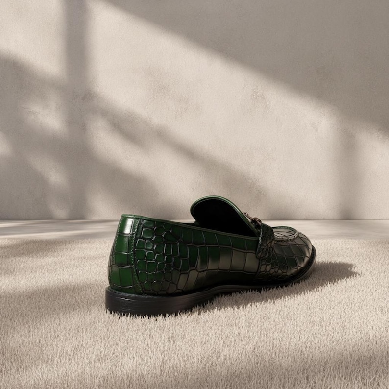 Puede incluir: Un mocas&iacute;n de cuero verde oscuro con textura de cocodrilo. El zapato tiene una punta redondeada y un tac&oacute;n peque&ntilde;o. El mocas&iacute;n est&aacute; sobre una superficie clara.