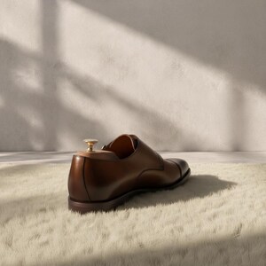 Side profile of a handmade cognac patina double monk strap shoe with clean cap toe, two gold buckle straps, dark leather outsole, low stacked block heel, and rich hand-burnished cowhide upper — bespoke men's formal dress shoe.