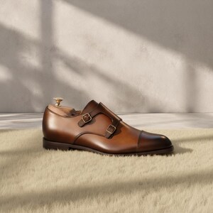 Close-up of the double gold-tone monk strap buckles and hand-burnished cognac patina on a genuine cowhide leather dress shoe, showing warm amber-to-chocolate tonal gradation, cap toe seam stitching, and strap construction detail.