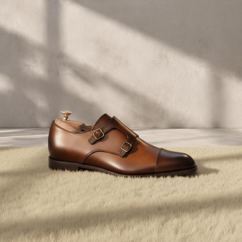 Close-up of the double gold-tone monk strap buckles and hand-burnished cognac patina on a genuine cowhide leather dress shoe, showing warm amber-to-chocolate tonal gradation, cap toe seam stitching, and strap construction detail.
