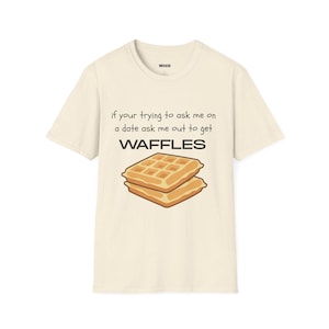 Puede incluir: Una camiseta de manga corta de color crema con textura moteada. El frente presenta un gráfico de dos gofres dorados apilados y el texto en negro: if your trying to ask me on a date ask me out to get WAFFLES. El logotipo de la marca wcco está impreso en el interior.