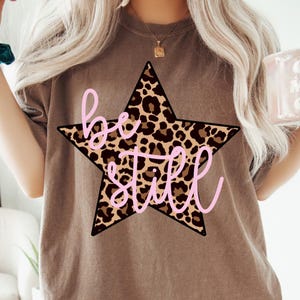 May include: Brown t-shirt featuring a star design with a leopard print pattern. The star has a black outline and the words "be still" in pink cursive. The shirt is a neutral color.