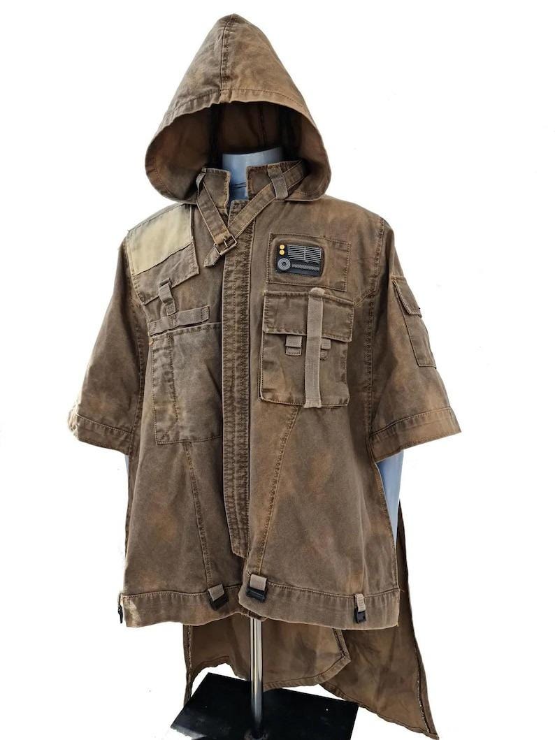 May include: A brown hooded jacket with multiple pockets and a unique design. The jacket has a short sleeve design and a long, flowing tail. It features a patch with a small rectangular design. The jacket is displayed on a mannequin.