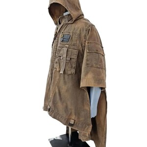 May include: Brown hooded jacket with multiple pockets and a patch detail. The jacket has a unique design with a long, flowing back panel. The garment is displayed on a mannequin, showcasing its style and construction. The jacket appears to be made of a durable, textured fabric.