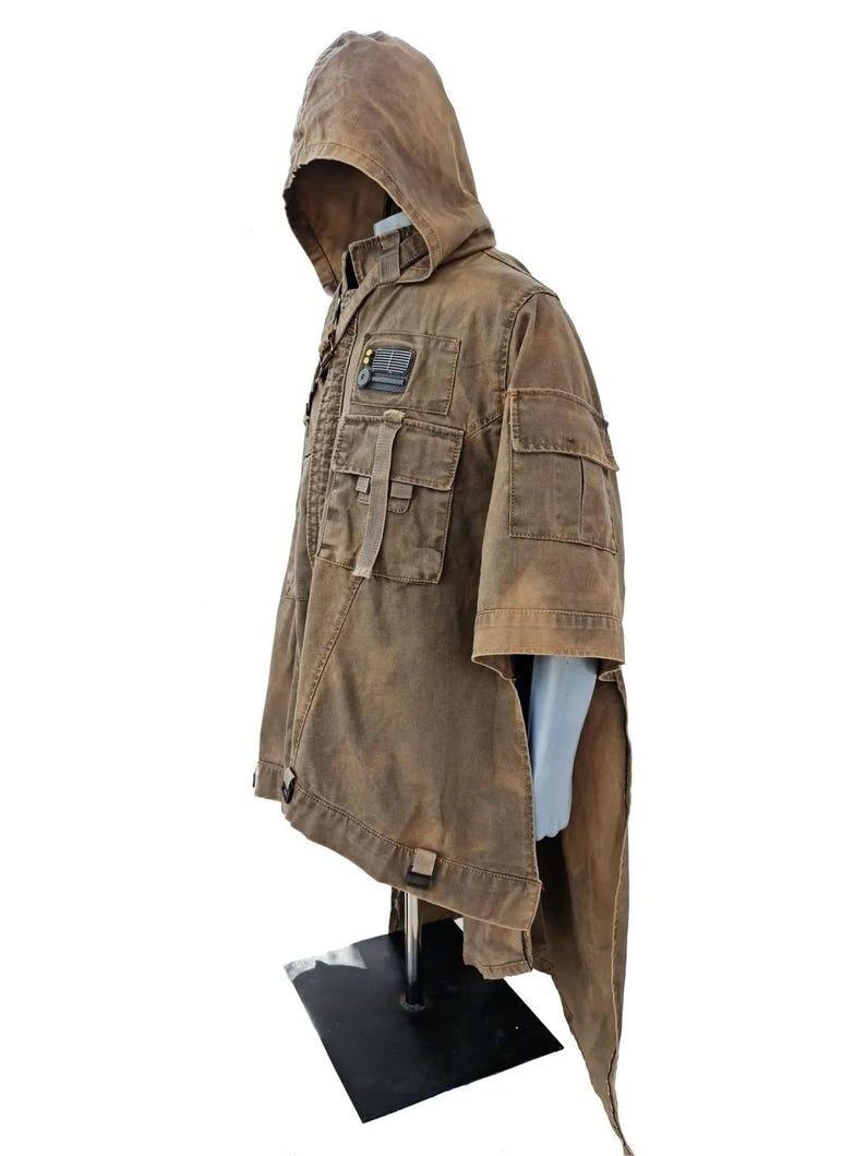 May include: Brown hooded jacket with multiple pockets and a patch detail. The jacket has a unique design with a long, flowing back panel. The garment is displayed on a mannequin, showcasing its style and construction. The jacket appears to be made of a durable, textured fabric.