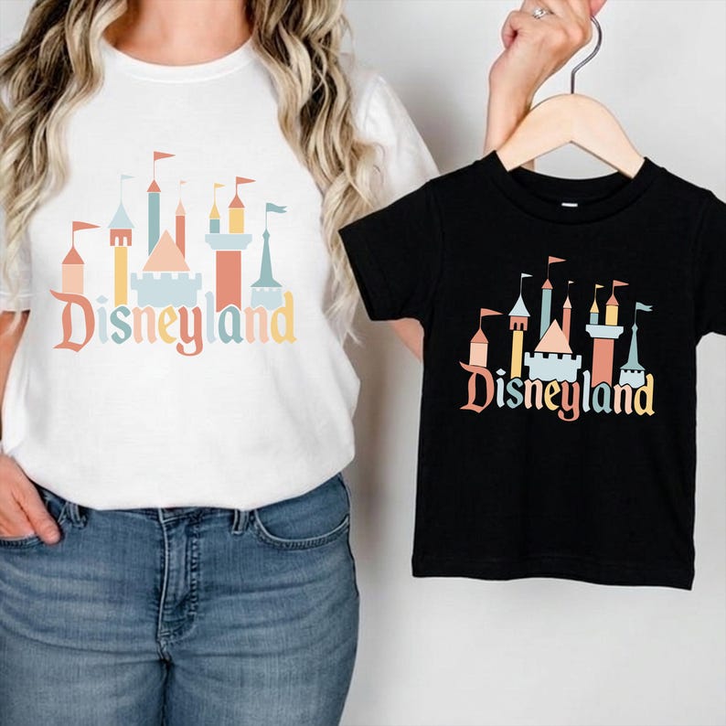 A woman wearing a white t-shirt with a Disneyland castle graphic and holding a small black t-shirt with a similar graphic.