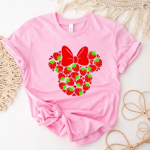 A pink t-shirt with a red bow and a design of strawberries is displayed on a white surface, accompanied by a woven mat and some straw accessories.