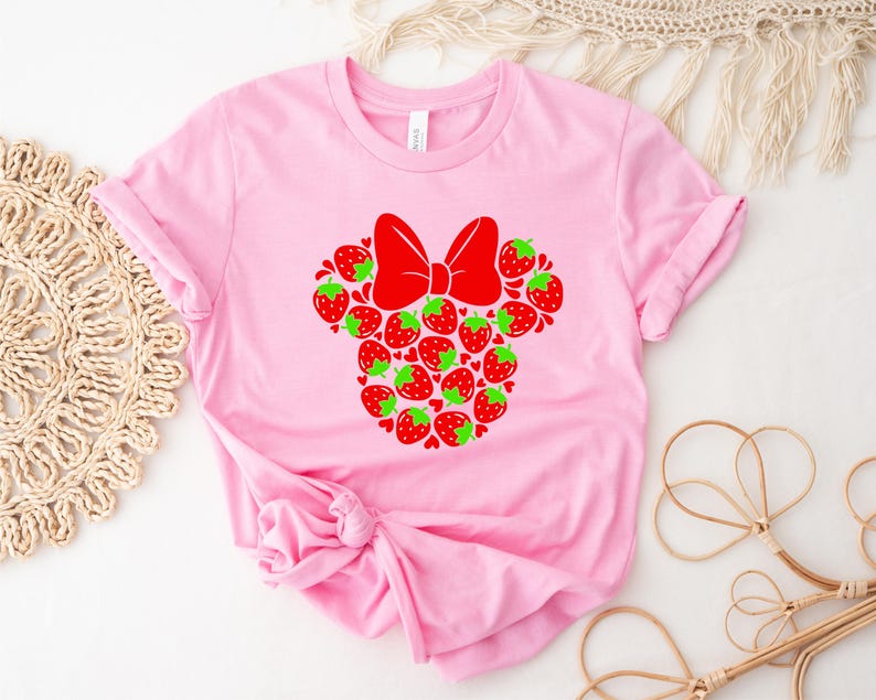 A pink t-shirt with a red bow and a design of strawberries is displayed on a white surface, accompanied by a woven mat and some straw accessories.