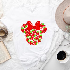 a white t-shirt with a red bow design, a hat, a book, a cup of coffee, and some other items arranged on a white surface.