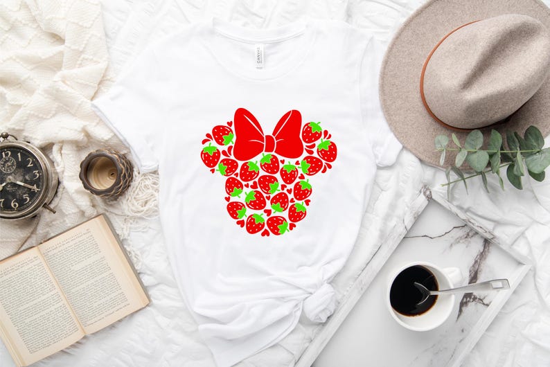 a white t-shirt with a red bow design, a hat, a book, a cup of coffee, and some other items arranged on a white surface.