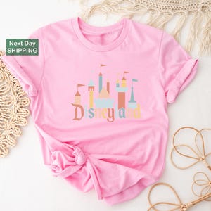A pink t-shirt with a Disney-themed graphic design, including a castle and other iconic landmarks, is displayed on a white background.