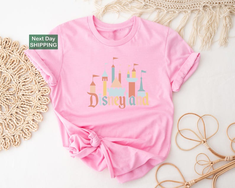 A pink t-shirt with a Disney-themed graphic design, including a castle and other iconic landmarks, is displayed on a white background.