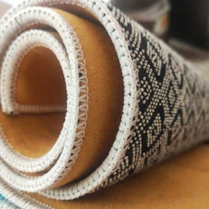 K&ouml;nnte beinhalten: Aufgerollter Teppich mit einem geometrischen schwarz-wei&szlig;en Muster auf der obersten Schicht. Das Innere des Teppichs ist einfarbig senfgelb, und die Kanten sind mit einem wei&szlig;en, gen&auml;hten Rand versehen.