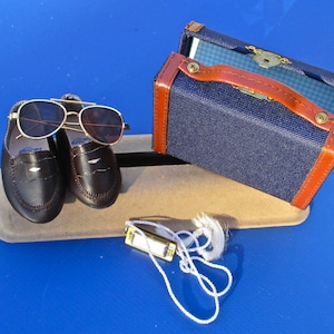 May include: A small blue suitcase with brown trim, a pair of brown leather loafers, a pair of aviator sunglasses, and a gold-coloured harmonica with a white string attached.