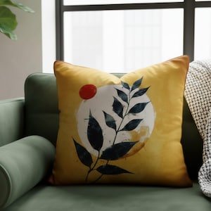 May include: A square decorative pillow with a yellow background and a design featuring a white circle, a red dot, and dark blue leaves. The pillow has a brown border and is placed on a green sofa.