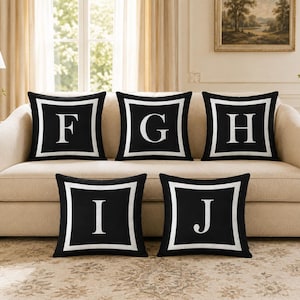 Op de afbeelding: Een set van vijf vierkante sierkussens in zwart met een witte rand en een grote witte letter in het midden van elk kussen. De letters F, G, H, I en J zijn weergegeven in een schreeflettertype. De kussens zijn in een groep gerangschikt, met drie op de bovenste rij en twee op de onderste rij.