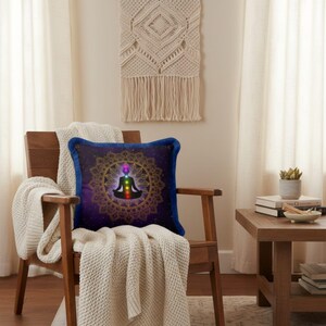 May include: A square decorative pillow with a blue fringe features a meditating figure with chakra symbols inside a gold mandala design. The pillow is on a wooden chair with a cream-colored blanket.