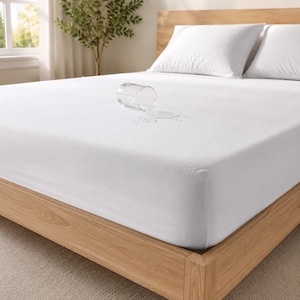 May include: A white mattress protector fitted over a bed, featuring a smooth, solid-colored surface. A clear glass of water is tipped over on the mattress protector, with a small puddle of spilled water visible on the white fabric surface.