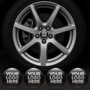 May include: A silver alloy car wheel with a black tire. The wheel has a star-like design with five spokes. The center cap displays the text "YOUR LOGO HERE" in white on a black background. Four additional caps are below.