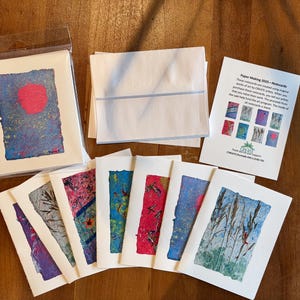 May include: A collection of handmade notecards with abstract art designs. The cards feature a variety of colors, including blue, red, and purple. The image also includes white envelopes and a promotional card with the text "Paper Making 2023 - Notecards."