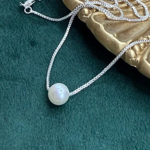 May include: A delicate silver necklace with a single, luminous white pearl pendant. The chain has a fine, box-style design, and the pearl is centered, creating a minimalist and elegant look. The necklace is displayed on a green velvet surface.