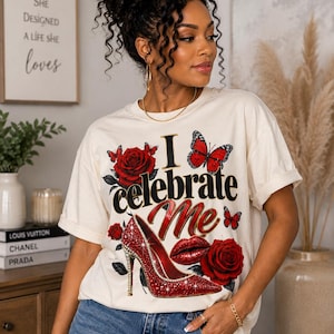 Può includere: Una t-shirt a maniche corte color crema con una stampa grafica che riporta il testo I celebrate Me, una scarpa con tacco rossa ricoperta di glitter, rose rosse, labbra rosse e farfalle rosse. La t-shirt è abbinata a un paio di jeans blu.