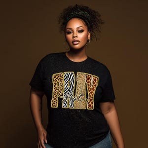 Könnte beinhalten: Schwarzes Glitzer-T-Shirt mit den Worten "PRAY Just" in goldener Umrandung. Die Buchstaben sind mit Tiermustern gefüllt: Leopard, Zebra und Giraffe. Das Shirt hat einen Rundhalsausschnitt.