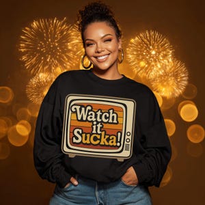 Op de afbeelding: Zwart sweatshirt met een retro televisie-afbeelding met de tekst "Watch it Sucka!". De afbeelding heeft oranje, gele en bruine strepen. De persoon die het sweatshirt draagt, lacht en draagt oorringen.