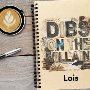May include: A spiral-bound notebook with the text "DIBS ON THE VILLAIN" in a decorative font, with the name "Lois" below. A cup of coffee with latte art and a pen are on a wooden surface.