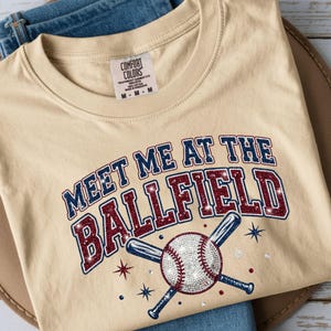May include: Cream-colored t-shirt with the text "MEET ME AT THE BALLFIELD" in navy blue and red glitter lettering. A baseball and crossed bats graphic is below the text. The shirt is folded on a brown hat.
