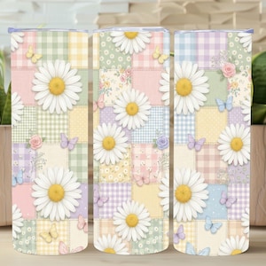 Puede incluir: Tres vasos de acero inoxidable con un diseño de margarita y patchwork. El diseño presenta grandes margaritas blancas con centros amarillos, mariposas y pequeñas rosas sobre un fondo de patchwork pastel.
